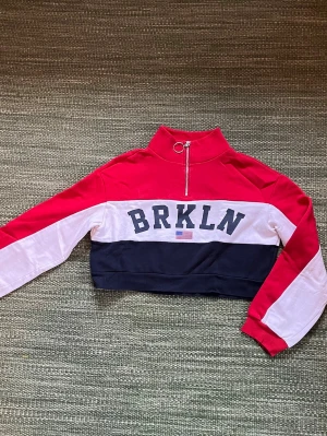 Collagetröja BRKLN Divided H&M XS - Säljer en  långärmad collagetröja från Divided H&M i storlek XS. Tröjan har färgblock i rött, vitt och mörkblått, BRKLN-tryck framtill och en cool half zip med ringdetalj. Perfekt till jeans eller kjol för en sportig vibe.