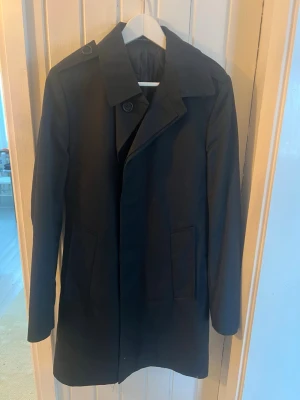 Svart trenchcoat från Tiger of Sweden - Säljer en svart trenchcoat från Tiger of Sweden i klassisk modell med bälte i midjan och knappar. Jackan har quiltat foder och två stora fickor framtill. Perfekt för dig som vill ha en stilren och tidlös look.