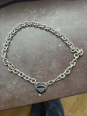 Tiffany & Co halsband - Klassisk kedja i äkta silver från Tiffany & Co med hjärtformat hänge där det står 'Please return to Tiffany & Co New York 925'. Kedjan har grova länkar och ett robust lås, perfekt för att ge din outfit en lyxig touch.