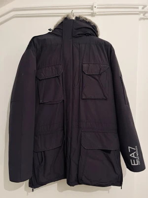 Svart parkajacka EA7 Emporio Armani - Säljer en mörkgrå dunjacka från EA7 Emporio Armani med stor huva och päls runt kanten. Jackan har flera stora fickor framtill, patch med logga på ärmen och dragkedja. Perfekt för kalla dagar och riktigt snygg streetstil. Dragkedjor på sidorna gör det möjligt att justera storleken.