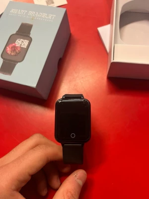 Smart Bracelet - Smart Bracelet med rektangulär pekskärm och svart silikonarmband. Funktioner för pulsmätning och blodtryck, enligt förpackningen. Klockan ser ut att vara i gott skick utan synliga repor eller skador. Levereras med originalförpackning.