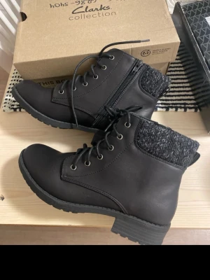 Svarta boots från Clarks med snörning - Säljer ett par svarta boots från Clarks med snörning och dragkedja på sidan. Skorna har en ribbad, stickad kant upptill i grått och svart samt en robust platt sula. Tillverkade i syntetmaterial med rund tå och låg klack. Perfekta för höst och vinter!