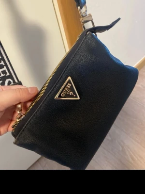 Svart axelväska från Guess med logorem - Säljer en svart axelväska från Guess i syntetmaterial med guldfärgade detaljer och dragkedja. Väskan har bred axelrem med Guess-logga och en liten avtagbar miniväska på remmen. Invändigt finns flera fack och dragkedjeförslutning. Perfekt för dig som vill ha både stil och funktion.