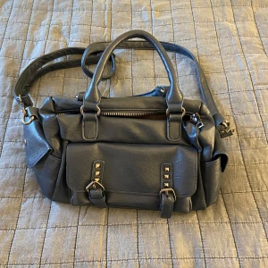 Mörkblå axelväska med nitar - Snygg mörkblå axelväska i skinnimitation med både handtag och justerbar axelrem. Väskan har flera fack, dragkedjor och en framficka med coola nitar och spännen. Perfekt storlek för allt du behöver ha med dig och passar till många olika stilar.