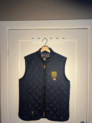 Marinblå quiltad dunväst Morris - Säljer en marinblå quiltad dunväst från Morris med gul broderad logga och brittisk flagga på bröstet. Västen har dragkedja framtill, ståkrage och två fickor. Perfekt för lager-på-lager och snygg till både skjorta och hoodie.
