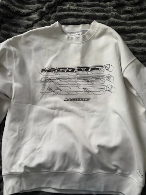 Vit sweatshirt från Lacoste - Säljer en vit sweatshirt från Lacoste med coolt grafiskt tryck på bröstet och klassisk rund hals. Tröjan har långa ärmar och en relaxed passform, perfekt för en chill och stilren look. Liten Lacoste-logga nertill på framsidan.