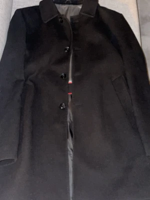 Svart kappa från Italian Job, ull/kashmir - Stilren svart kappa från Italian Job i storlek 52. Tillverkad i en lyxig mix av ull och kashmir med slät yta och klassisk krage. Kappan har knäppning framtill, diskreta sidofickor och helfodrad insida med snygga detaljer.