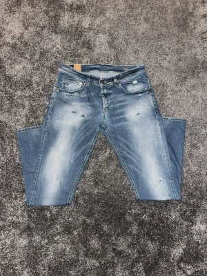 Blå Dondup jeans med slitningar  - Säljer ett par blå Dondups jeans med slitningar och ljusa partier framtill och baktill. Klassisk femficksmodell med knappgylf och snygga detaljer på bakfickorna. Jeansen har en slim-fit passform och är tillverkade i skönt jeansmaterial.