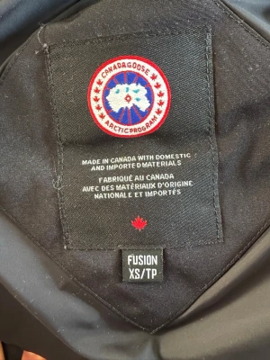 Svart dunjacka från Canada Goose XS - Säljer en svart Canada Goose dunjacka i storlek XS. Jackan har en fluffig pälsdetalj på huvan. Det är varm och skön och är perfekt i dessa tider även över snygg 