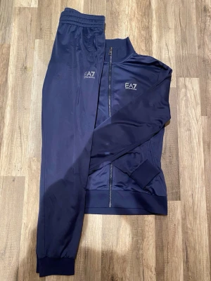 Marinblå EA7 tracksuit - Säljer en marinblå tracksuit från EA7 Emporio Armani. Koftan har hög krage, hel dragkedja framtill och logga på bröstet. Tillverkad i ett glansigt syntetmaterial som känns sportigt och modernt. Perfekt för dig som gillar stilrena och bekväma plagg.