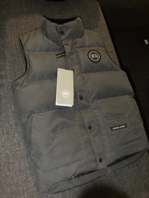 Svart dunväst från Canada Goose - Snygg svart dunväst från Canada Goose med hög krage, två stora framfickor och klassisk logotyp på bröstet. Västen har tryckknappar och är tillverkad i slitstarkt material med dunfyllning för extra värme. Perfekt för lager-på-lager och streetwear.