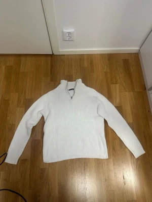 Vit stickad tröja med halv dragkedja - Klassisk vit stickad tröja med halv dragkedja vid halsen. Tröjan har lång ärm och ribbstickade muddar vid ärmslut och nederkant. Perfekt för lager på lager och enkel att matcha med olika outfits.