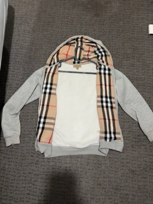 Grå hoodie med Burberry-mönstrad huva - Snygg grå hoodie från Burberry med klassiskt beige, svart och rött rutmönster på insidan av huvan. Tröjan har långa ärmar, ribbade muddar och en avslappnad passform. Perfekt för dig som gillar stilrena och lyxiga detaljer.