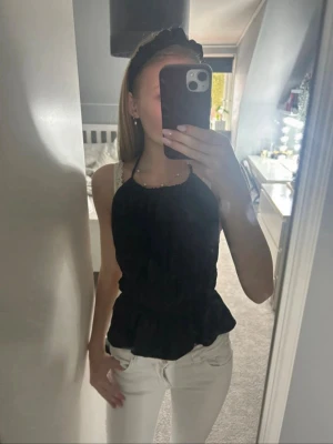 Svart halterneck topp med knyt - Snygg svart halterneck topp med knyt i nacken och midjan. Toppen har en lätt volang nertill och är perfekt för en trendig look. Materialet känns lätt och luftigt, och modellen är croppad med en smickrande passform.