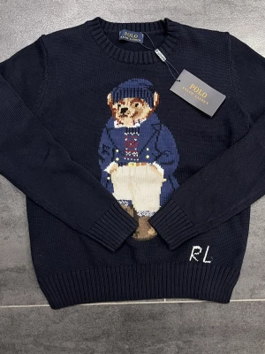 Mörkblå stickad tröja Polo Bear - Mörkblå stickad tröja från Polo Ralph Lauren med den ikoniska Polo Bear på bröstet. Tröjan har rund halsringning, ribbade muddar och RL-broderi nedtill. Perfekt för dig som gillar klassisk preppy stil och vill ha något soprent men ändå lekfullt.