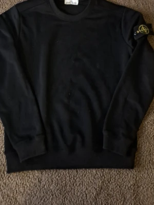 Svart stickad tröja från Stone Island - Säljer en svart stickad tröja från Stone Island med klassisk rund hals och ribbade muddar. Tröjan har den ikoniska Stone Island-loggan på vänster ärm. Perfekt för dig som gillar stilrena och tidlösa plagg.