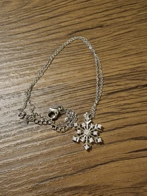 Silverfärgat halsband med snöflinga - Snyggt halsband i silverfärg med ett hängsmycke format som en snöflinga. Kedjan är tunn och smidig med ett klassiskt klolås. Pläterad. Längd på kedja 40 cm + 5 cm. Berlock 1,5 × 1,5 cm. Snöflingan har små glittrande stenar som ger extra bling till din look. Perfekt accessoar för dig som gillar vinterinspirerade smycken.