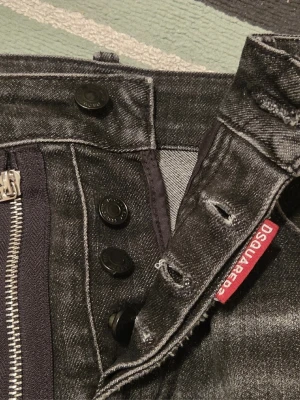 Svarta Dsquared2 jeans med dragkedjedetalj - Säljer ett par svarta Dsquared2 jeans med slitningar, vita färgstänk och en stor silverfärgad dragkedja med ringdetalj framtill. Jeansen har knappgylf, klassisk femficksmodell och Dsquared2-logga på röd tag vid fickan. Snygg tvätt och smal passform.
