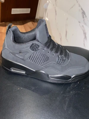 Svarta Nike Jordan 4 sneakers - Säljer ett par helsvarta Nike Jordan 4 sneakers med klassisk meshpanel på sidan, robust sula och snygga detaljer i syntetmaterial. Skorna har snörning och en sportig, chunky siluett som passar perfekt till streetwear.