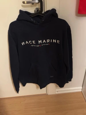 Mörkblå hoodie från Race Marine - Snygg mörkblå hoodie från Race Marine med vit text och koordinater på bröstet. Klassisk känguruficka framtill och huva med dragsko. Tillverkad i mjukt bomullsmaterial som är skönt att ha på sig. Perfekt för en avslappnad och sportig stil.