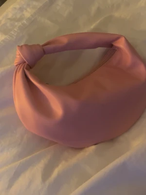 Rosa handväska med knutdetalj - Trendig rosa handväska i syntetmaterial med mjuk, rundad form och en unik knutdetalj på handtaget. Väskan har dragkedja upptill för säker förvaring och en stilren look som passar till många outfits. Sen här väskan är verkligen så snygg till sommaren jag säljer den då jag har för många väskor 💕