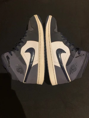 Nike Air Jordan 1 Mid Blå/Vit - Snygga Nike Air Jordan 1 Mid sneakers i marinblått och vitt. Skorna har klassisk snörning, vit och mörk blå sula och den ikoniska Nike Swoosh-loggan i mörkblått på sidan. Tillverkade i en mix av skinn och syntetmaterial för en clean och sportig look.