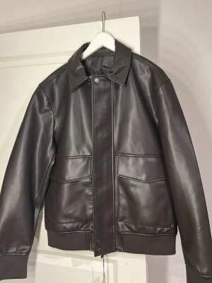 Brun skinnjacka med fickor - Snygg mörkbrun skinnjacka med klassisk krage och stora framfickor. Jackan har dragkedja och tryckknappar framtill samt ribbade muddar vid ärmslut och nederkant. Perfekt för dig som vill ha en stilren och tidlös look.