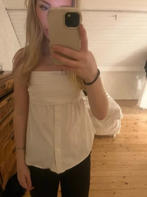 Vit axelbandslös topp med knappar - Säljer en vit axelbandslös topp med knäppning framtill och lös passform. Toppen har en söt volangdetalj upptill och är perfekt för varma dagar. Passar till både jeans och kjol. Har sytt in från en M till S /xs