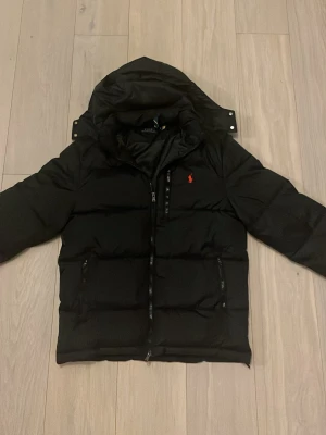 Svart pufferjacka från Polo Ralph Lauren - Säljer en svart pufferjacka från Polo Ralph Lauren med huva och dragkedja framtill. Jackan har det klassiska röda Polo-logot på bröstet och två sidofickor med dragkedja. Perfekt för kalla dagar och riktigt snygg till streetwear-stilen. Pris kan sänkas vid snabb affär