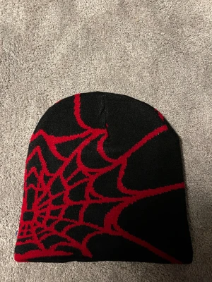 Svart mössa med rött spindelnätsmönster - Cool svart mössa med ett stort rött spindelnätsmönster som täcker hela mössan. Perfekt för dig som gillar unika accessoarer med lite edge. Materialet känns mjukt och stretchigt, troligen akryl eller bomullsblandning. Ingen synlig logga eller märke. Helt ny alldrig använd 