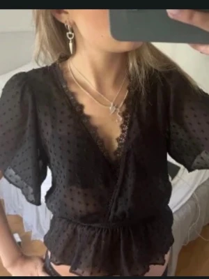 Svart blus med spets - Snygg svart blus i transparent material med prickig struktur och volangdetaljer. Djup v-ringning med spetskant och resår i midjan för en feminin siluett. Korta, luftiga ärmar. Perfekt att styla med smycken för en trendig look. dock med ett hål