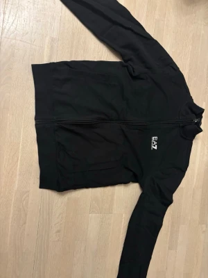 Svart ziptröja från EA7 Emporio Armani - Svart ziptröja från EA7 Emporio Armani med vit logga på bröstet och diskreta vita ränder i nacken. Tröjan har hög krage, hel dragkedja och två fickor framtill. Perfekt för en sportig och stilren look. Som ny 