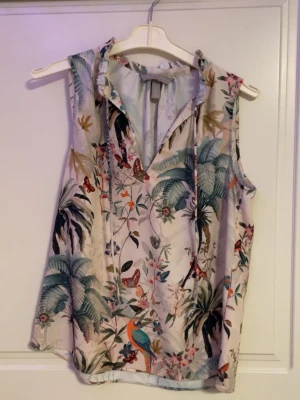 Blommig ärmlös blus med tropiskt mönster - Snygg ärmlös blus med tropiskt blommönster i grönt, blått och beige. Blusen har krage med knytband och är tillverkad i ett lätt och svalt material, perfekt för varma dagar. Färgglada detaljer med fåglar och fjärilar ger en unik look.