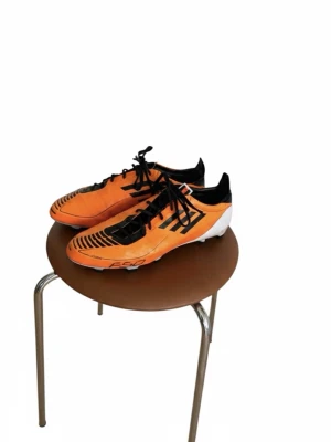 Orangea Adidas fotbollsskor F50 - Säljer ett par grymma orangea Adidas F50 fotbollsskor med svarta detaljer och snörning. Skorna har en låg profil och är designade för fart på planen. Ovandelen är i syntetmaterial och sulan är anpassad för spel på gräs.