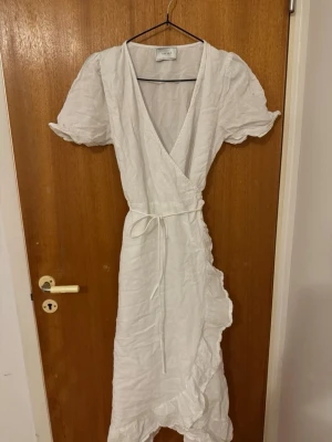 Vit omlottklänning med volang - Säljer en vit omlottklänning från Neo Noir med puffärmar, knyt i midjan och volangdetaljer längs nederkanten. Klänningen har v-ringning och är tillverkad i ett lätt linnematerial, perfekt för varma dagar.