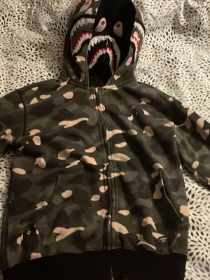 BAPE camo hoodie med shark-tryck - Säljer en svartvit hoodie från BAPE med ikoniskt shark-tryck på huvan och färgglada WGM-bokstäver på sidan. Dragkedja framtill, två huvor och ribbade muddar. Materialet är mjukt och bekvämt, köptes för 1500kr