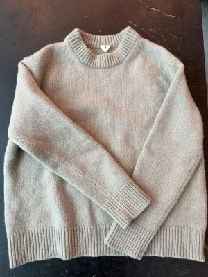 Beige stickad tröja med rund hals - Mysig beige stickad tröja med rund halsringning och ribbade muddar vid ärmslut och nederkant. Tröjan har en avslappnad passform och är perfekt för lager på lager. Materialet ser ut att vara mjukt och värmande, troligen ull eller ullmix.