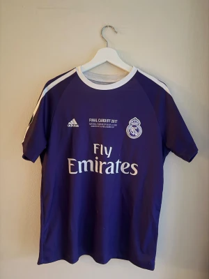 Real Madrid Ronaldo t-shirt Adidas - Säljer en lila Real Madrid t-shirt från Adidas med tryck för Champions League-finalen 2017 i Cardiff. Tröjan har korta ärmar, vita detaljer och texten 'Fly Emirates' på framsidan samt 'Ronaldo 7' på ryggen. Perfekt för dig som älskar fotboll och Real Madrid.