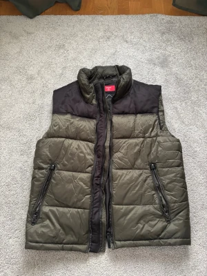 Olivgrön dunväst med svarta detaljer - Snygg olivgrön dunväst med svarta axelpartier och dragkedjor. Västen har hög krage och två praktiska fickor med dragkedja framtill. Perfekt för lager-på-lager och chill stil. Materialet är quiltat och ger en modern look.                                             Bara att höra av sig vid eventuella frågor!