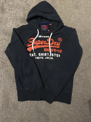 Superdry hoodie - Svart hoodie från Superdry med stort rött och vitt tryck på bröstet och japanska tecken. Klassisk huva med vita dragsnören och mjukt material. Perfekt för en avslappnad streetwear-look. Storlek L.
