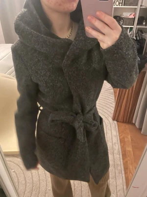 Mörkgrå unik kappa med bälte och stor krage - Snygg unik mörkgrå kappa med stor, mysig krage och knytskärp i midjan. Kappan har lång ärm och en klassisk, rak passform. Perfekt för kyliga dagar och enkel att styla till olika outfits. Passar även M. Använd gärna köp nu