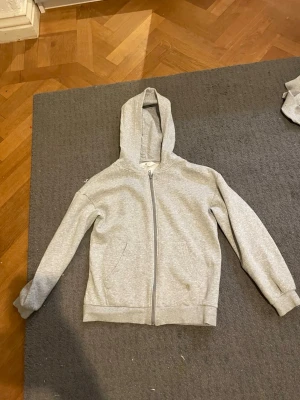 Ljusgrå zip hoodie med huva - En enkel och stilren ljusgrå hoodie med dragkedja framtill och två fickor. Hoodien har ribbade muddar vid ärmslut och nederkant samt en klassisk huva. Perfekt för chill dagar eller när du vill ha en clean look. Materialet känns mjukt och bekvämt.