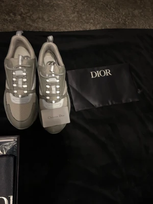 Gråa Dior B22 med dustbag och box - Pris kan diskuteras vid snabb affär.!!! Snygga sneakers från Dior i grått med vita detaljer. Skorna har en modern design med meshpaneler för extra andningsförmåga klassisk snörning framtill och diskret Dior-logga på sidan. Perfekta för dig som vill ha en exklusiv touch på din outfit.
