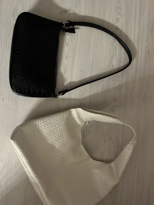 Svart och vit axelväska i crocopräglat - Två stilrena axelväskor, en i svart och en i vitt, båda med crocopräglat mönster. Den svarta har en klassisk, avlång form och smal axelrem medan den vita är större och mjukare i modellen. Perfekta för att lyfta vilken outfit som helst. Säljer båda tillsammans, man kan köpa enskild oxå