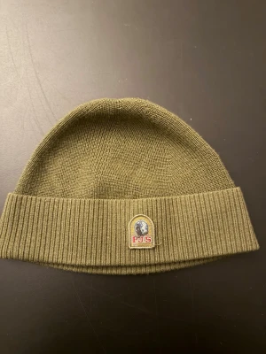 Olivgrön mössa från Parajumpers - Snygg olivgrön ribbstickad mössa från Parajumpers med uppvikt kant och klassisk PJS-logga framtill. Perfekt accessoar för att hålla värmen och ge en stilren touch till din outfit. Materialet är mjukt och stretchigt för skön passform. Köpt för 600 kr på kidsbrandstore