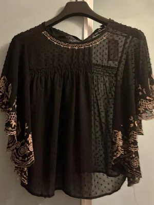 Svart blus med broderade detaljer - Svart blus i luftig mesh med prickigt mönster och vida korta ärmar. Ärmarna har vackra broderier i beige och vågig kant. Blusen har rund halsringning med broderad kant och är lätt transparent. Perfekt för en trendig och bohemisk look. Köpte den från sellpy, aldrig andvänd.