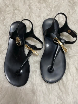 Svarta sandaler med rosett från Ted Baker - Stilrena svarta sandaler från Ted Baker med guldfärgade detaljer och en elegant rosett framtill. Tillverkade i syntetmaterial och har öppen tå samt platt sula. Perfekta för sommaren och ger en chic touch till din outfit.