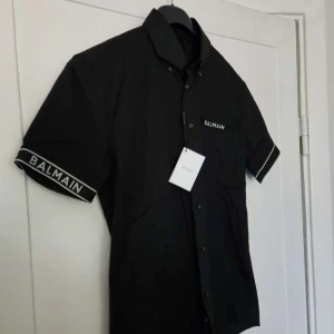 Svart kortärmad skjorta från Balmain - Svart skjorta från Balmain med korta ärmar och vit logotyp på ärmslutet. Säljer åt min bror då den nt kommer till användning! Har fortfarande kvar prislappen. (Pris kan diskuteras) står i dirham då den är köpt i Dubai och den är äkta, ordinarie pris 1,360 (dirham) vkt motsvarar 3500kr 