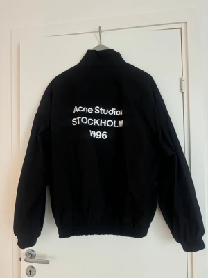 Svart vindjacka Acne Studios - Säljer en svart vindjacka från Acne Studios med vit text på ryggen där det står 'Acne Studios STOCKHOLM 1996'. Jackan har hög krage, dragkedja framtill och resår vid ärmslut och midja. Perfekt för dig som gillar minimalistisk och stilren design. Nästan aldrig använd. Skulle säga att den även passar M 