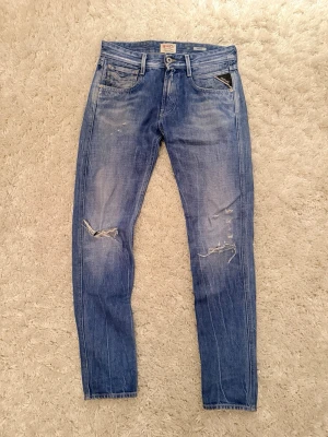 Blå slitna jeans från Replay - Säljer ett par blå jeans från Replay med slitningar och hål på knäna. Modellen har smal passform, klassisk femficksdesign och detaljerade sömmar. Jeansen har en tvättad look och är tillverkade i bomull med lite stretch för extra komfort.